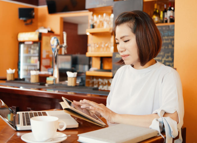 Asian female café owner sitting at a counter, looking stressed while reviewing finances on a tablet and laptop, symbolising small business debt challenges.
